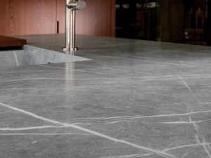 Neolith Zaha Stone from the MGD brands collection - a close-cropped, low level view of a grey stone worktop with white veining. In the blurred background you can make out a modern sink and mixer tap in brushed steel.