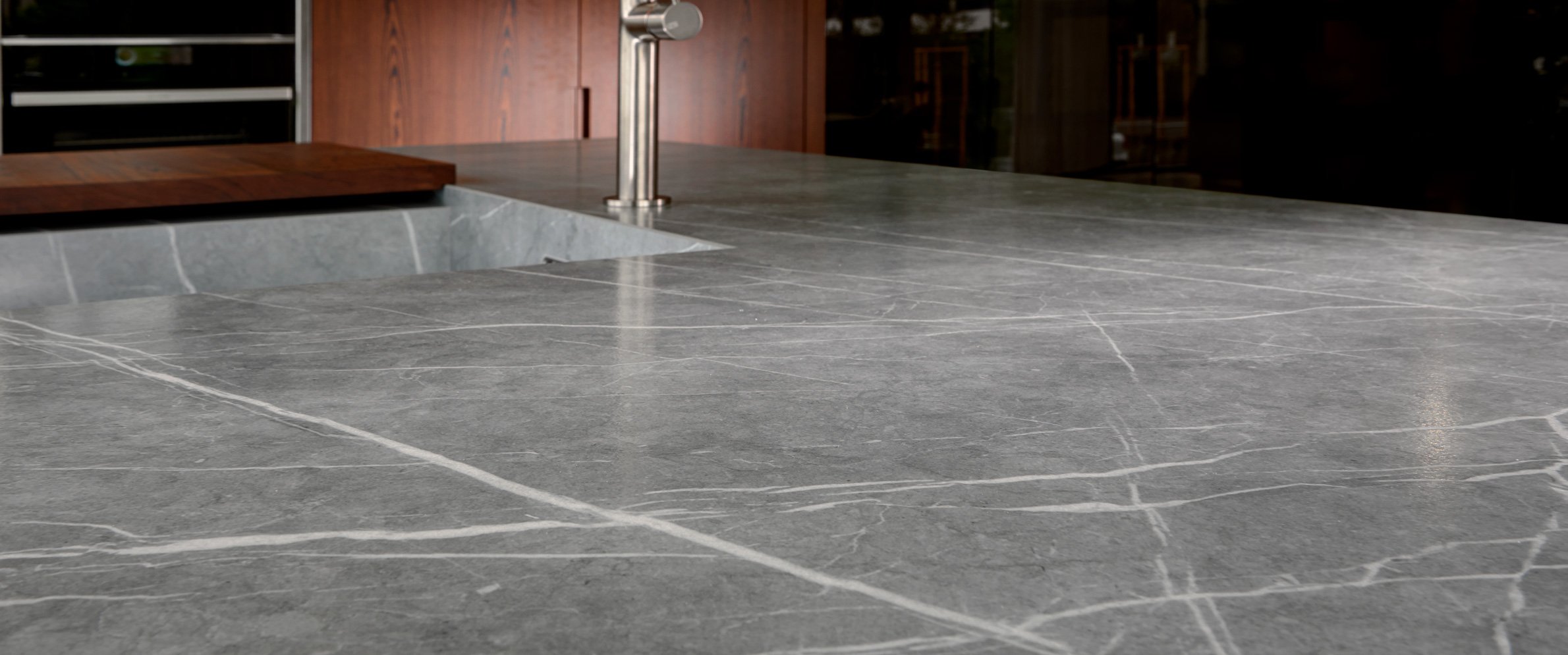 Neolith Zaha Stone from the MGD brands collection - a close-cropped, low level view of a grey stone worktop with white veining. In the blurred background you can make out a modern sink and mixer tap in brushed steel.