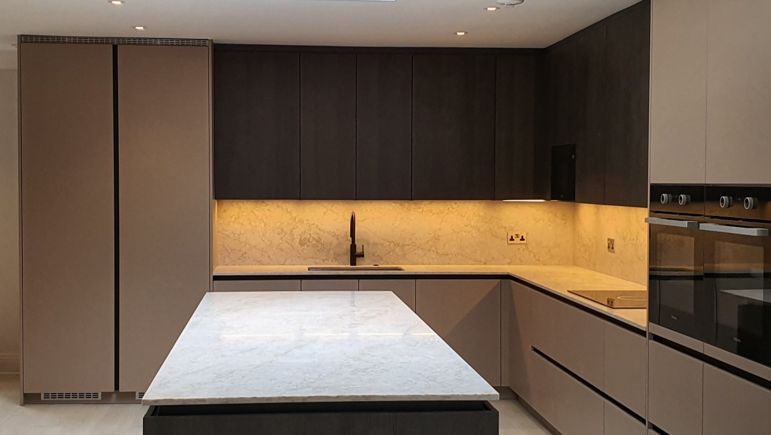 Technistone Quartz Herefordshire - kitchen installation with white marble kitchen island and worktops, with splashbacks in the background. dark coloured kitchen cabinets contrast the marble look and muted yellow downlighting creates ambience across the background