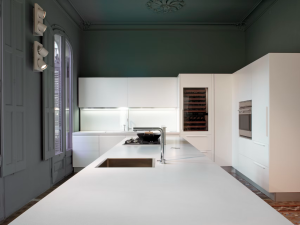 Modern kitchen with green walls, white cabinets, stainless steel appliances, and a wine cooler built into the wall. The kitchen island has a white quartz Blanco Zeus countertop and stainless steel sink. Decorative moulding and a painted ceiling add architectural interest to the space.