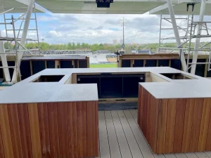 The final image provides a broader view of the servery area, highlighting its open and accessible layout. The countertops form a U-shape, maximizing the serving area while maintaining a streamlined look. The background reveals a stunning view of the cricket ground, enhancing the bar's appeal as a prime location for guests.